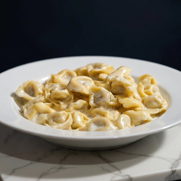 A white bowl filled with creamy tortellini pasta, sprinkled with black pepper, on a marble surface.